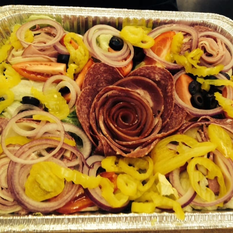Antipasto salad with salami, ham, onions, tomatoes, olives, and banana peppers in a foil pan.