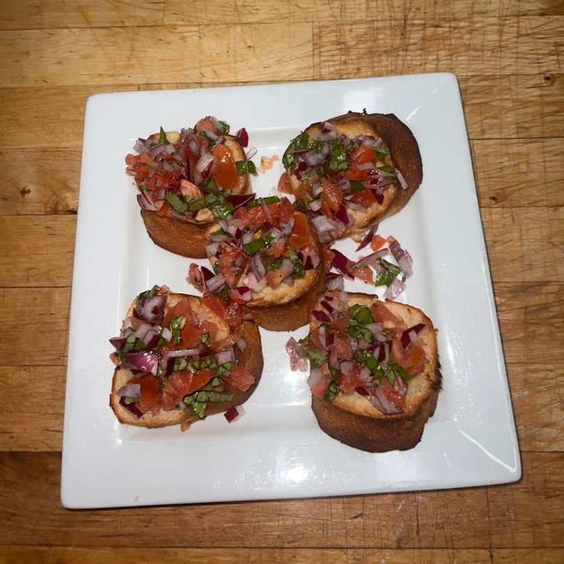 Four bruschetta appetizers with diced tomatoes, red onions, and basil on a white rectangular plate.